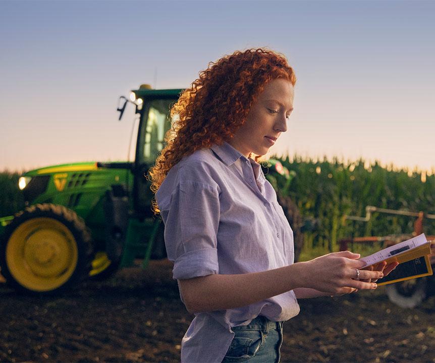 Woman with booklet stands in front of tractor in cane field. 